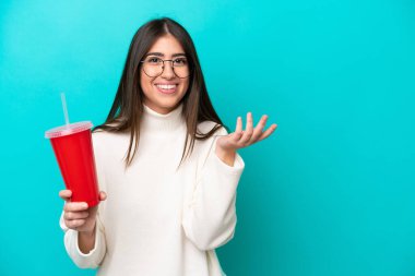 Mavi arka planda izole edilmiş gazoz içen genç beyaz kadın davet etmek için elini yana uzatıyor.