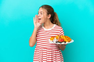 Little caucasian girl holding waffles isolated on blue background shouting with mouth wide open to the side