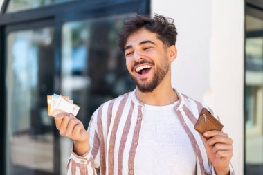 Açık havada yakışıklı bir Arap 'ın elinde cüzdanı ve mutlu bir ifadesi var.