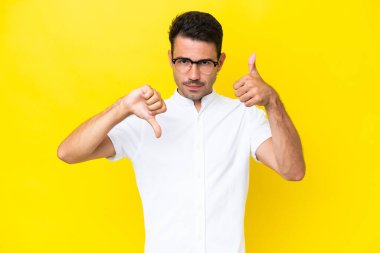 Soyutlanmış sarı arka planda genç yakışıklı adam iyiye-kötüye işaret yapıyor. Evet ya da hayır arasında kararsız