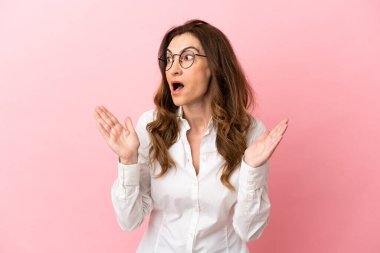 Orta yaşlı beyaz kadın, pembe arka planda yalıtılmış, şaşırmış bir yüz ifadesi var.