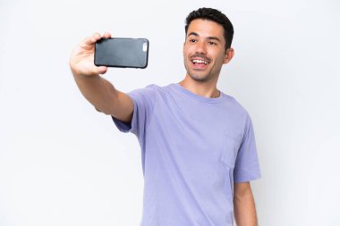 İzole edilmiş beyaz arka planda genç yakışıklı bir adam cep telefonuyla selfie çekiyor.