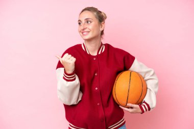 Pembe arka planda izole edilmiş genç basketbolcu kadın bir ürünü sunmak için kenarı işaret ediyor.
