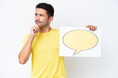 İzole edilmiş beyaz arka plan üzerinde genç yakışıklı bir adam konuşma baloncuğu ikonlu bir levha tutuyor ve düşünüyor