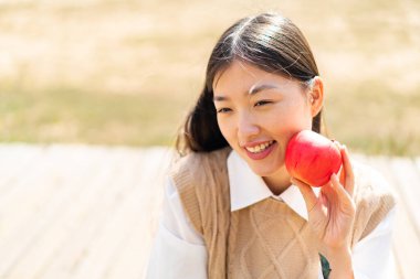 Açık havada genç bir Çinli kadın elinde mutlu bir ifadeyle bir elma tutuyor.