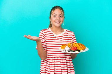 Little caucasian girl holding waffles isolated on blue background with shocked facial expression