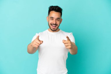 Mavi arka planda izole edilmiş genç beyaz adam şaşırmış ve ön tarafı işaret ediyor.