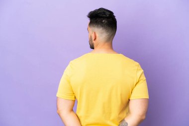 Young caucasian man isolated on purple background in back position and looking back