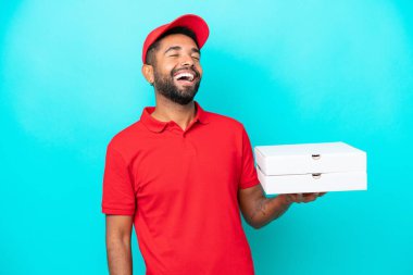 Pizza teslimatı üniformalı Brezilyalı adam mavi arka planda izole edilmiş pizza kutuları alıyor ve gülüyor.