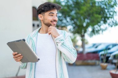Handsome Arab man holding a tablet at outdoors thinking an idea and looking side