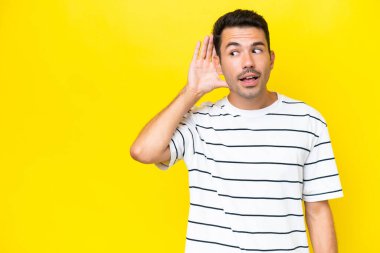 Izole sarı arka plan üzerinde genç yakışıklı adam kulak elini koyarak bir şey dinlerken