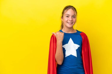 Sarı arka planda izole edilmiş Süper Kahraman küçük kız bir ürünü sunmak için kenarı işaret ediyor.