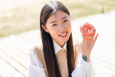 Young Chinese woman at outdoors holding a donut and happy