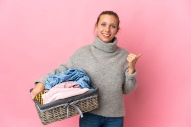 Elinde pembe arka planda izole edilmiş bir çamaşır sepeti olan genç bir kadın bir ürünü sunmak için kenarı işaret ediyor.