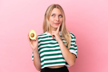 Young caucasian woman holding an avocado isolated on pink background having doubts while looking up