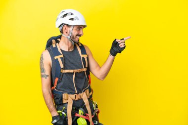 Genç beyaz dağcı sarı zemin üzerinde izole edilmiş bir şekilde parmak göstererek bir ürün sunuyor.
