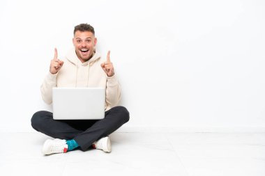 Young caucasian man with a laptop sitting on the floor isolated on white background pointing up a great idea