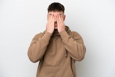 Beyaz arka planda izole edilmiş beyaz tenli yakışıklı adam gözlerini elleriyle kapatıyor.