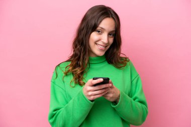 Pembe arka planda izole edilmiş genç beyaz kadın cep telefonuyla bir mesaj gönderiyor.