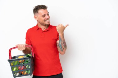 Beyaz arka planda izole edilmiş yiyecek dolu bir sepet taşıyan genç beyaz bir adam bir ürün sunmak için kenarı işaret ediyor.