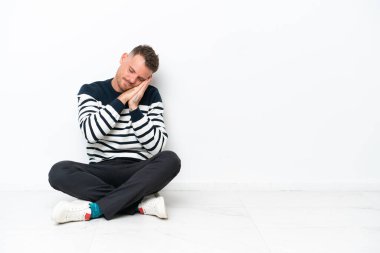 Yerde oturan genç adam, beyaz arka planda izole bir şekilde uyku jesti yapıyor.