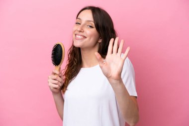 Young caucasian woman with hair comb isolated on pink background saluting with hand with happy expression