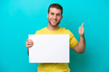 Young caucasian man isolated on blue background holding an empty placard with thumb up