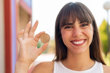 Açık havada elinde mutlu bir ifadeyle Bitcoin tutan genç bir kadın.