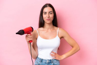 Young caucasian woman holding a hairdryer isolated on pink background with sad expression