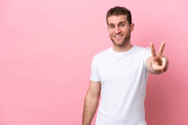 Pembe arka planda izole edilmiş genç beyaz adam gülümsüyor ve zafer işareti gösteriyor.