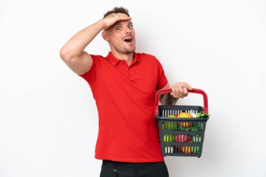 Young caucasian man holding a shopping basket full of food isolated on white background doing surprise gesture while looking to the side