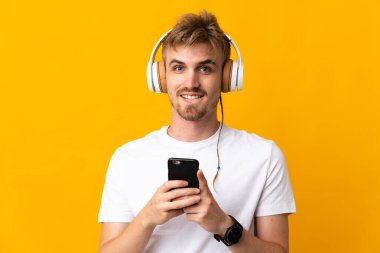 Young handsome blonde man isolated on yellow background listening music with a mobile and looking front
