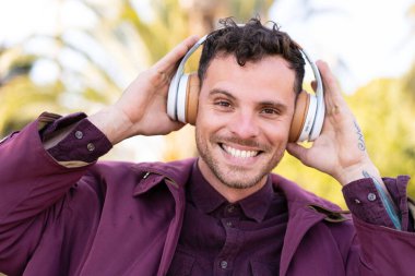 Genç beyaz bir adam açık havada müzik dinliyor.