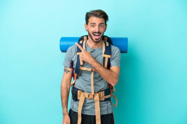 Mavi arka planda izole edilmiş büyük sırt çantalı genç dağcı. Şaşırmış ve işaret ediyor.