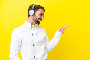 Tele pazarlamacı beyaz adam sarı arka planda izole edilmiş bir kulaklıkla çalışıyor. Parmakla göstererek bir ürün sunuyor.