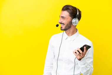 Tele pazarlamacı beyaz adam sarı arka planda izole edilmiş bir kulaklıkla çalışıyor cep telefonuyla sohbet ediyor.