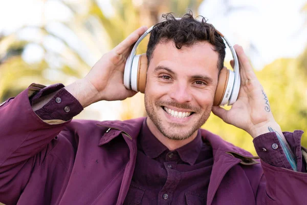 Genç beyaz bir adam açık havada müzik dinliyor.