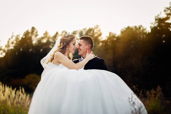 Great portrait of a wedding couple, which is walking in the field. A groom holding his bride on his hands, while she is embracing him and smiling.