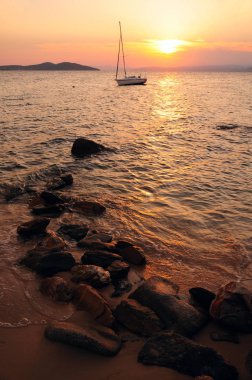 Bir tekne ve ön planda taşlarla denizde gün batımı, Yunanistan, Halkidiki, Ouranoupoli yakın