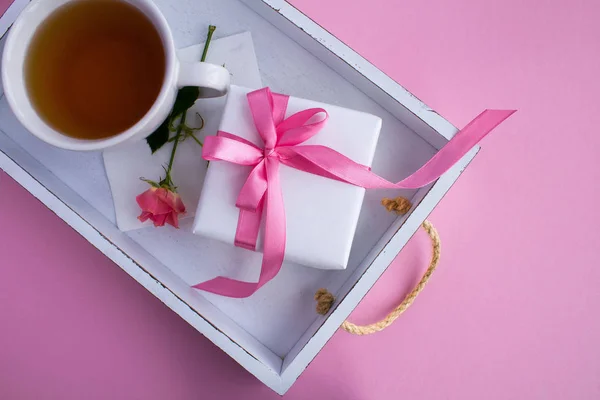 Gift with pink bow and tea in the cup on the white wooden tray on the light pink  background.Top view.Closeup.