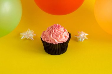 Sarı zemin üzerine pembe krema ve renkli balonlarla bir cupcake