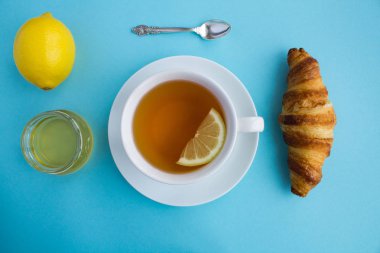 Çay saati. Bir fincan sıcak çay, kruvasan, bal ve limon mavi zemin üzerine. Üstten görünüm. Kopya alanı.