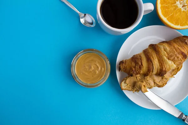 Breakfast: croissant with peanut paste,black coffee and orange on the blue background.Top view.Copy space.