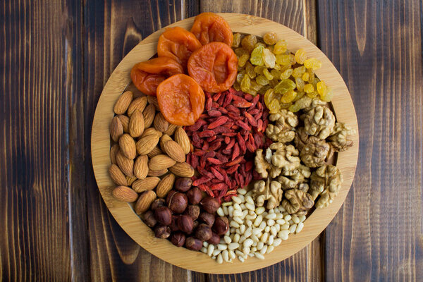 Dried apricots, raisins, goji berries ,different nuts on the round cutting board on the brown  wooden background.Top view.