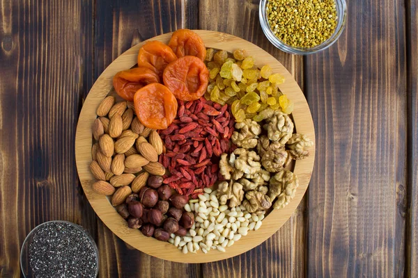 stock image Dried apricots, raisins, goji berries,different nuts, chia seeds and  bee pollen on the round cutting board on the brown  wooden background.Top view.