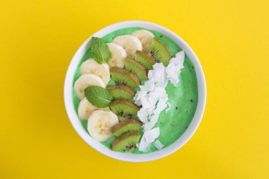 Sarı arka planın ortasında muz, kivi ve hindistan cevizi cipsi ile Smoothies kase. Üst te. Kopyalama alanı.