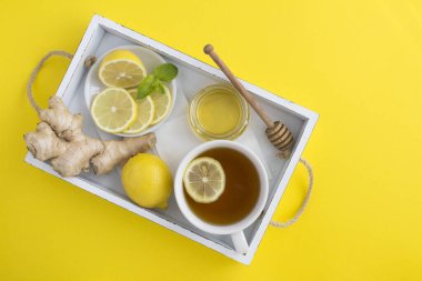 Sarı yüzeydeki beyaz ahşap tepside limonlu, ballı ve zencefilli yeşil çay manzarası.