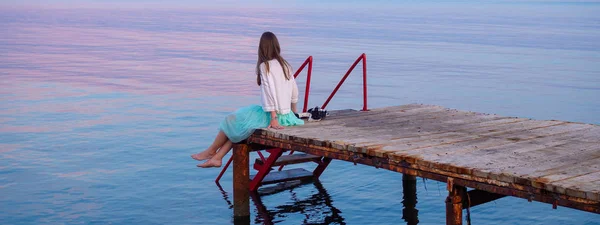 Side view of a wooden pier on the sea at sunset with young woman sitting on it, admirring amazing sunset, Durres, Albania. Awesome sunset sky