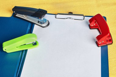 Two staplers and hole puncher lie over office folder with a spring clip or clipboard  and the blank of white  paper, with copy space, mockup for design. clipart