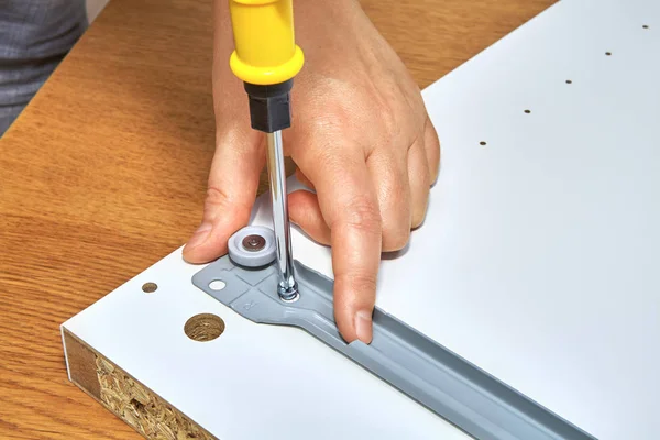 The handyman tightens the screw in drawer runner of wooden table with a screwdriver, flat pack furniture assembly.